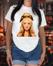 Load image into Gallery viewer, A woman wearing a white graphic t-shirt featuring a cartoon illustration of a blonde girl with a pumpkin-patterned bow, paired with denim shorts in a Halloween-themed room.

